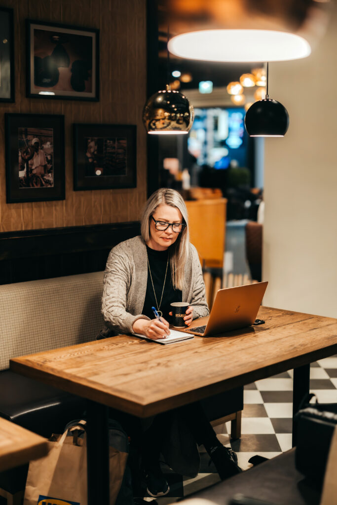 Ulkoistettu markkinointipäällikkö työskentelemässä kahvilassa kannettavan tietokoneen ja muistiinpanojen kanssa, suunnitellen ja johtaen yrityksen markkinointia kokonaisuutena.