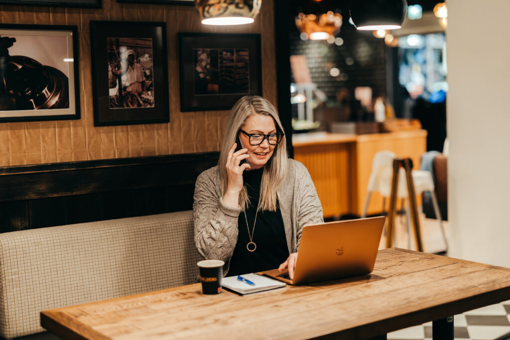 Markkinointia tukeva asiantuntija työskentelemässä kahvilassa kannettavan tietokoneen, muistivihon ja kahvikupin kanssa, tarjoten edullista markkinointiapua ja sparrausta aloitteleville yrittäjille.